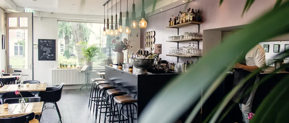 Bistro mit Theke und Barhockern im Vordergrund, dahinter Tische am Fenster.