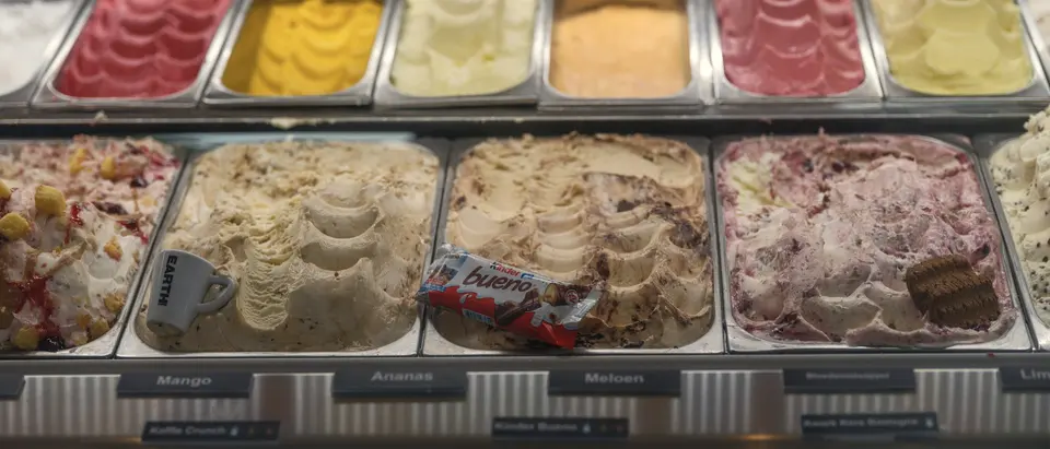 Eistheke mit zwei Reihen offener Behälter in Edelstahl, jede Sorte beschriftet.
